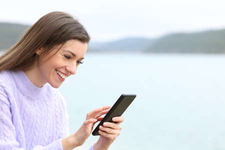 Happy teenager checking mobile phone content sitting in a lakeの写真素材