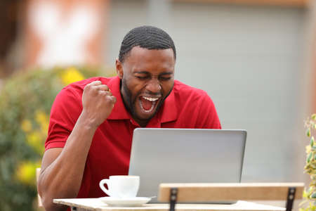 Excited man with black skin checking laptop content celebrating in a bar terraceの写真素材