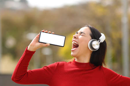 Happy woman in red wearing headphones singing in the street with blank smart phone screenの写真素材