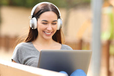 Happy woman using laptop sitting on bench with headphones in a parkの写真素材