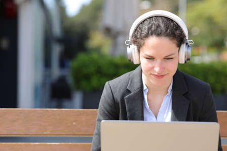Businesswoman wearing wireless headphones using laptop sitting on benchの写真素材