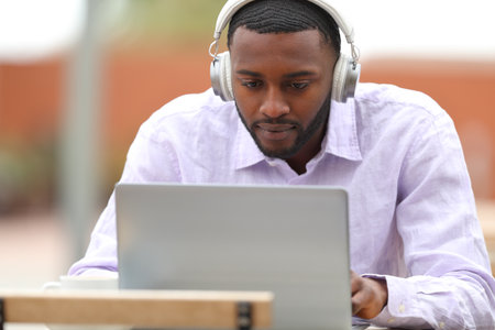 Front view portrait of a man with black skin using laptop with wireless headphones in a barの写真素材