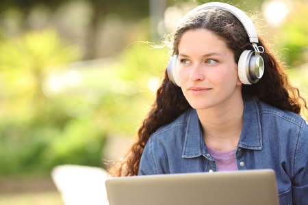 Woman with wireless headphones and laptop looks at side in a parkの写真素材