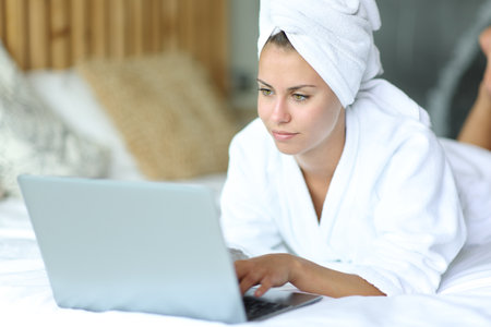 Beautiful woman lying on a bed after showering using laptop at homeの写真素材