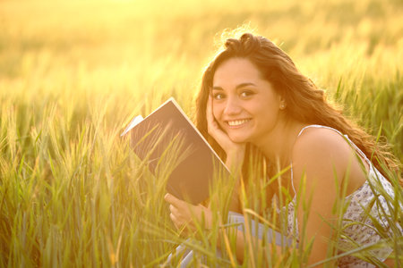 Happy reader holding book looks at camera in a wheat field at sunsetの写真素材