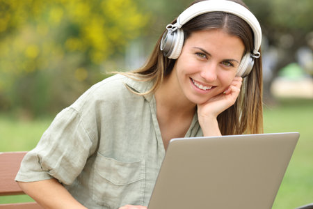 Happy teen with headphones and laptop looks at you in a parkの写真素材