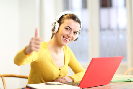 Happy student with headphones and laptop at home with thumbs up looking at you