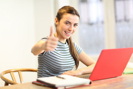 Happy student gesturing thumbs up at home with a laptopの写真素材
