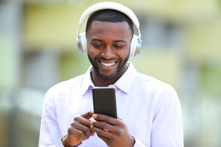 Front view portrait of a happy black man wearing wireless headphones listening to music online on phoneの写真素材