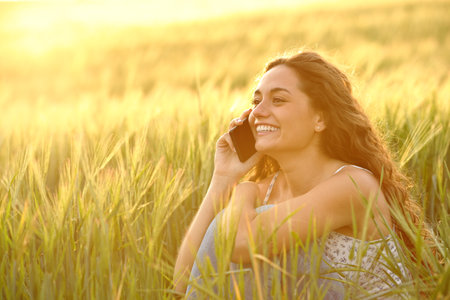 Happy woman talking on phone sitting in a field at sunriseの写真素材