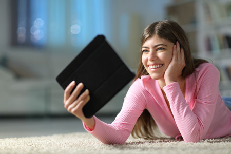 Happy teen watching media on tablet lying on the floor in the night at homeの写真素材