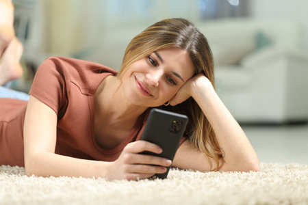 Woman checking phone lying on the carpet at homeの写真素材