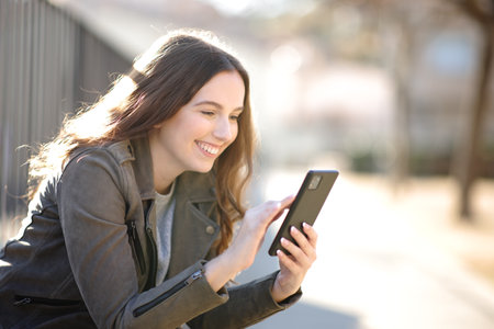 Happy woman using cell phone sitting in the street on a sunny dayの写真素材