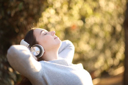 Relaxed woman wearing headphone resting listening audio sitting on a bench at sunset in a parkの写真素材
