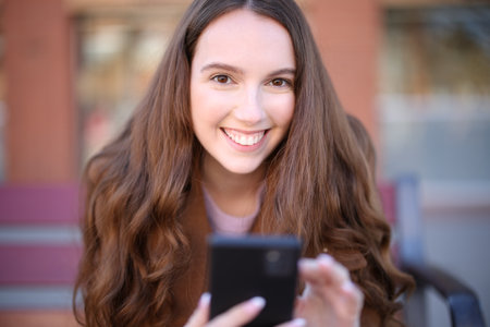 Front view of a happy woman on a bench looking at you holding phone in the streetの写真素材