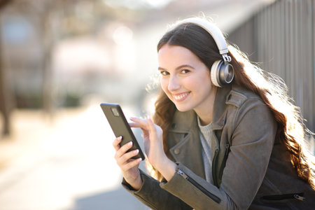 Happy woman wearing headphone looks at you listening audio with smart phone in the streetの写真素材