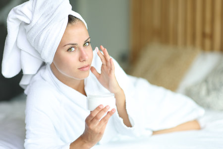 Woman hydrating face with moisturizer cream lying on a bed at home after showering looking at cameraの写真素材