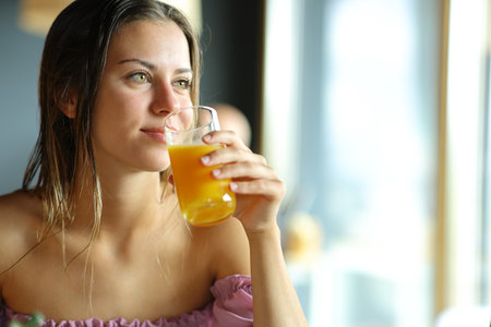 Happy woman drinking orange juice looking through a window in a restaurantの写真素材