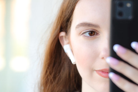 Front view close up portrait of a serious woman face wearing earbud using phone in the streetの写真素材