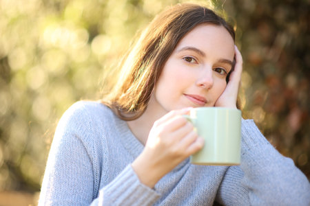 Pensive woman sitting in a park holding coffee looks at you at sunsetの写真素材