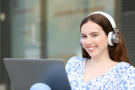 Happy woman wearing headphone posing looking at camera with laptop in the streetの写真素材