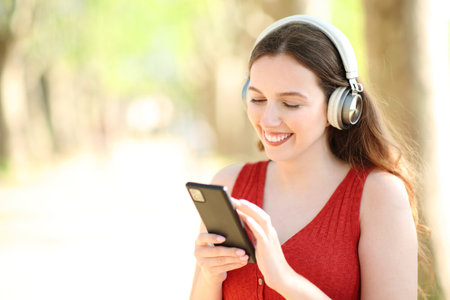 Happy woman wearing headphone listening to music using smart phone standing in a parkの写真素材