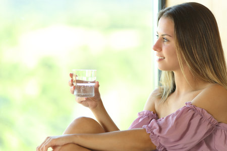 Happy teen holding glass of water looks through a window at home or hotelの写真素材