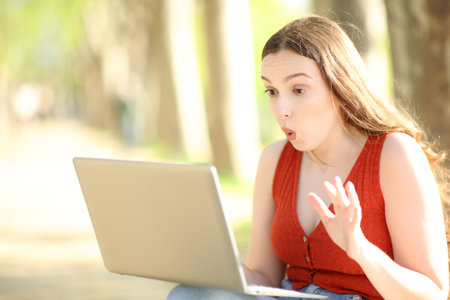 Surprised woman checking laptop content sitting alone in a parkの写真素材
