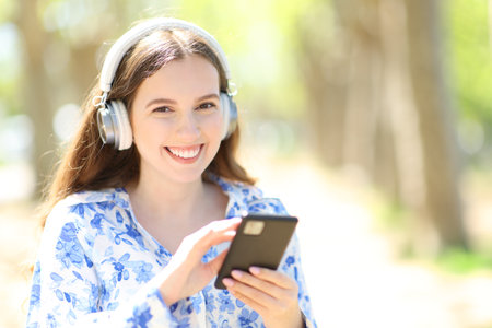Happy woman wearing headphone listening music holding smartphone looking at camera outdoorsの写真素材