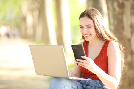 Happy woman using laptop and cell phone sitting in a park on summerの写真素材