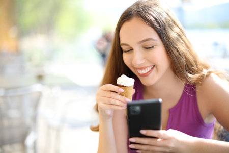 Happy woman eating ice cream and using phone sitting in a bar terraceの写真素材