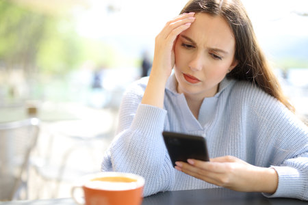 Worried woman checking phone sitting in a restaurant terrace on a sunny dayの写真素材