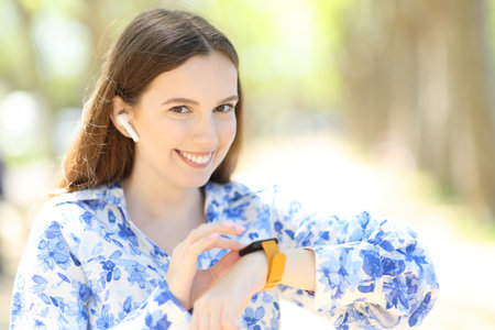 Happy woman looks at you checking music on smartwatch in a parkの写真素材
