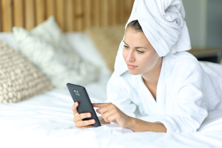 Satisfied woman using phone on bed after showeringの写真素材