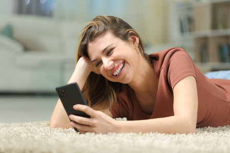 Happy woman watching phone videos lying on a carpet at homeの写真素材
