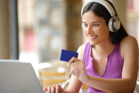 Happy woman wearing headphone buying online ppv media content with a laptop and credit card in a coffee shopの写真素材