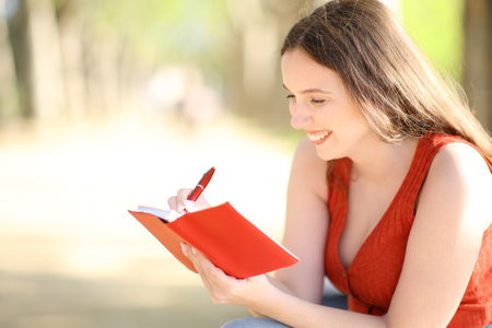 Happy woman writing in red agenda sitting alone in a parkの写真素材