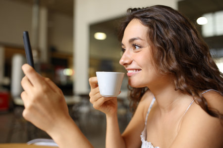 Happy woman drinking coffee using cell phone sitting in a barの写真素材