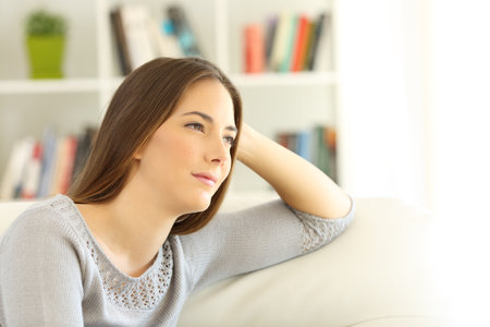 Portrait of a pensive woman looking away sitting on a couch at homeの写真素材