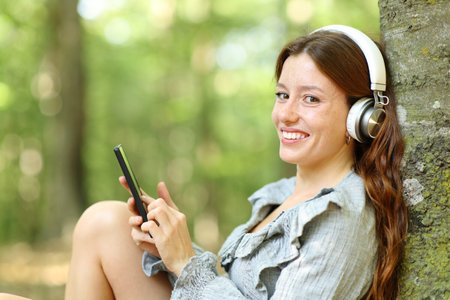 Happy woman listening to music with headphone and phone looks at you in a forestの写真素材