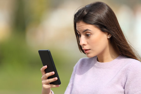 Shocked woman checking smart phone news in a parkの写真素材
