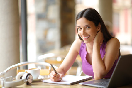 Happy student with a laptop posing looking at camera taking notes in a bar terraceの写真素材
