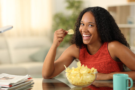Happy black woman eating potato chips looking at you at homeの写真素材
