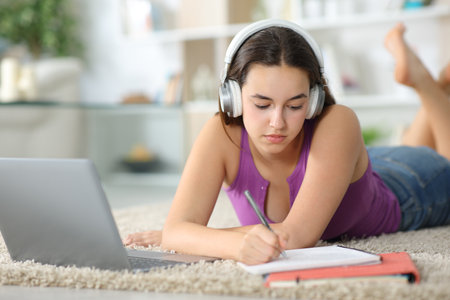 Happy student e-learning with laptop and headphone writing notes lying on the floor at homeの写真素材