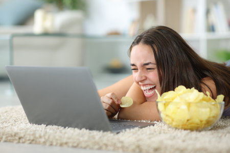 Funny woman watching media on laptop eating chips laughing hilariously at homeの写真素材