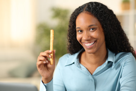 Happy black woman showing snack at camera at homeの写真素材