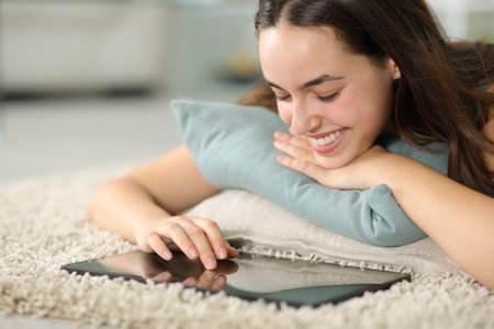 Happy woman using tablet lying on carpet at homeの写真素材