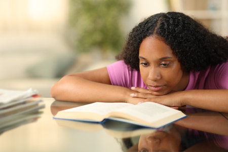 Serious black woman reading a paper book on a desk at homeの写真素材