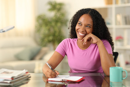 Happy black woman writing in agenda dreaming looking at side at homeの写真素材