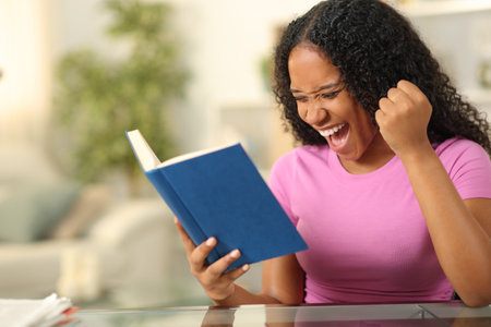 Black excited woman reading a paper book celebrating at homeの写真素材